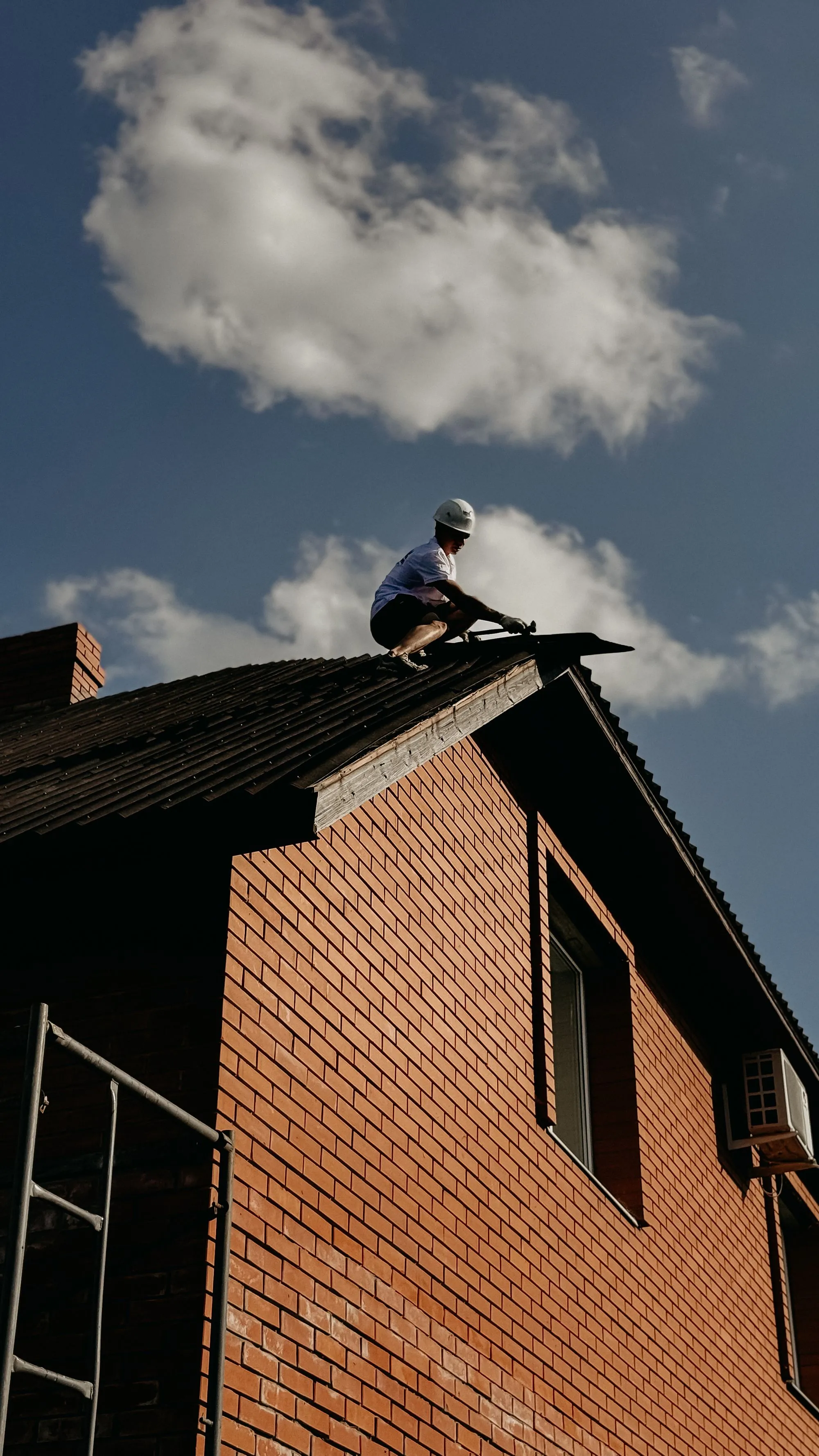 A roofer working along the ridge of a steep residential roof against the sky